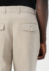 Pantalons ajustés beige clair avec poches à l'arrière, fermeture à bouton et texture fine. Accent de rayure subtile sur une poche.