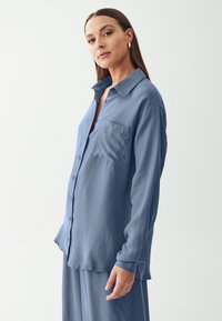 Camicia blu in satin a maniche lunghe con colletto a punta, chiusura a bottoni sul davanti e taschino sul petto, caratterizzata da una vestibilità rilassata e una superficie liscia.