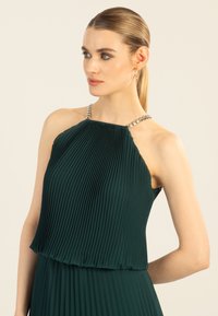 Grünes plissiertes Kleid mit einem Halterneck, ausgestattet mit einem dekorativen silbernen Besatz am Kragen. Glattes Material mit fließender Textur.