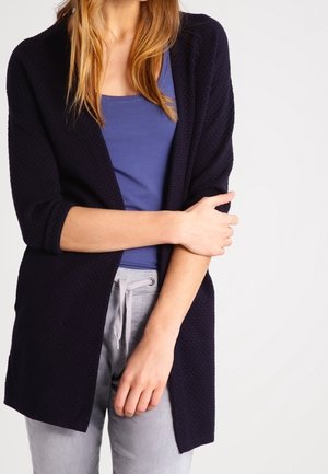 Mujer con camiseta sin mangas azul, cárdigan abierto texturizado negro y pantalones grises claros con cuerda, con los brazos cruzados frente a su torso.