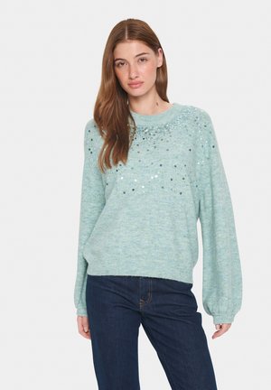 Suéter texturizado de color azul claro con cuello redondo, mangas abullonadas y detalles de lentejuelas en el pecho. Combinado con jeans de mezclilla oscura.