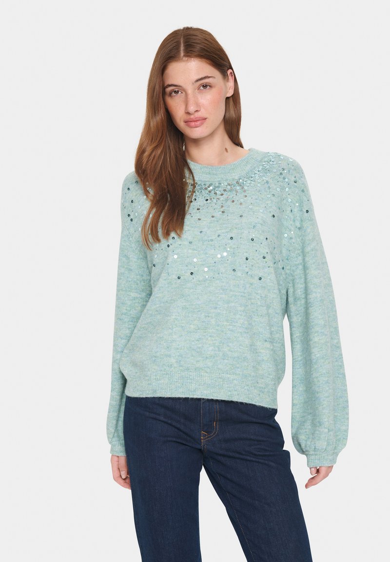 Ljusblå texturerad tröja med rund hals, ballongärmar och paljetter som detaljer över bröstet. Matchas med mörka denimjeans.