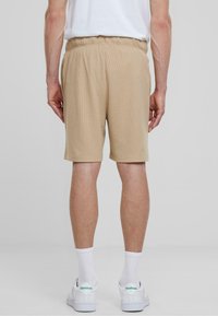 Beige, textúrájú, rugalmas derékú rövidnadrág, a comb közepéig ér. Fehér zoknikkal és zöld részletekkel ellátott sportcipővel viselik.