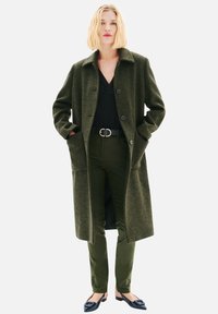 Manteau surdimensionné à carreaux verts avec col, deux poches avant et boutons, accompagné d'un pantalon vert foncé et de ballerines noires. Texture de tissu lisse.