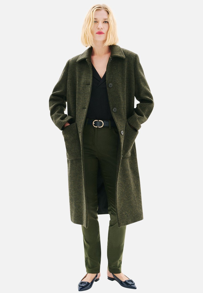 Manteau surdimensionné à carreaux verts avec col, deux poches avant et boutons, accompagné d'un pantalon vert foncé et de ballerines noires. Texture de tissu lisse.