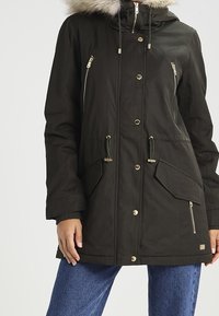 Parka noire avec capuche bordée de fourrure, boutons-pression à l'avant, poches zippées et à rabat, portée avec un jean bleu.