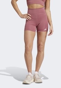 Kvinde iført mauve højtaljede træningsshorts og sports-BH med beige løbesko, stående med den ene hånd på hoften mod en ensfarvet baggrund.