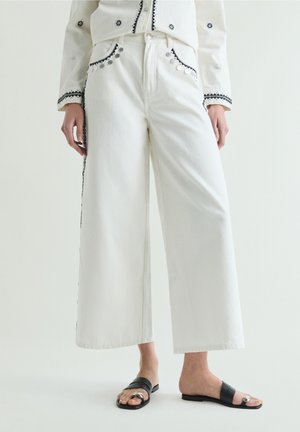 Pantalones blancos de pierna ancha con ribete decorativo negro y detalles de monedas plateadas, combinados con sandalias negras de punta abierta con detalle de hebilla plateada.