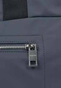 Sac à dos en tissu gris avec une fermeture éclair argentée dotée d'un tirette rectangulaire, accentué par une sangle texturée et des coutures soignées.