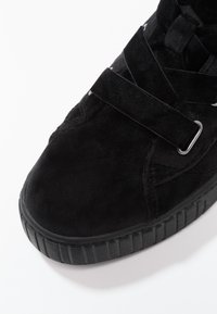 Mustat mokkanahkaiset urheilukengät, joissa on pyöreä kärki, paksu kumipohja, jossa on pystysuorat raidat, ja ristikkäiset hihnat, jotka on kiinnitetty metallisilla soljilla.