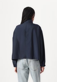 Giacca cropped blu navy in tessuto leggero con polsini elasticizzati e dettaglio a spacco sul retro. Abbinata a jeans in denim blu chiaro.