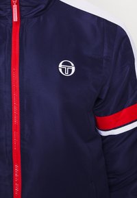 Marinejakke med rød glidelås og hvite detaljer. Har et rundt logo og rød stripe på ermene. Laget av glatt, teksturert materiale.