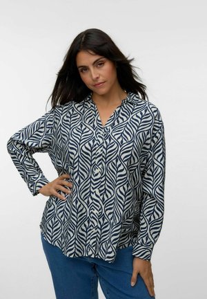 Vrouw met lang donker haar die een knoopjesoverhemd met lange mouwen en een blauw-wit bladmotief draagt, poseert met één hand op haar heup.