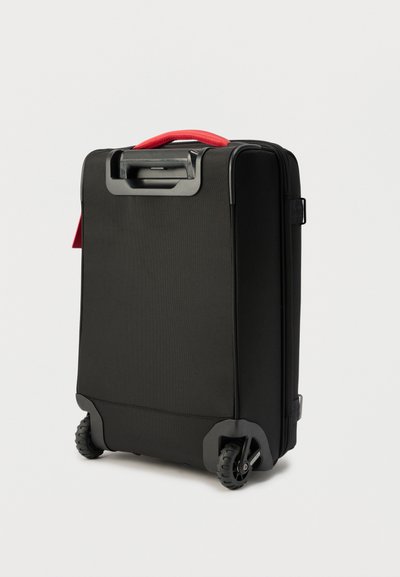 Μαύρη μαλακή βαλίτσα με δύο τροχούς, αναδιπλούμενο χερούλι και κόκκινο υφασμάτινο επάνω χερούλι σε λευκό φόντο.