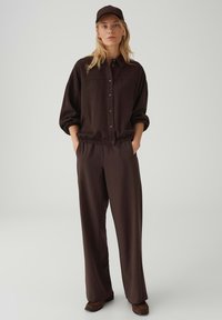 Donna che indossa una camicia marrone scuro con bottoni, pantaloni ampi abbinati, scarpe marroni e cappello marrone scuro, in piedi con le mani nelle tasche.