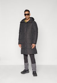 Doudoune noire avec capuche, présentant un design matelassé et une longueur longue. Portée sur un pantalon sombre et des chaussures noires, avec des lunettes de soleil.