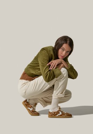 Giacca in velluto a coste verde, dettagli color crema, pantaloni bianchi, sneakers marroni con motivo e dettagli bianchi. Modello accovacciato su uno sfondo chiaro.