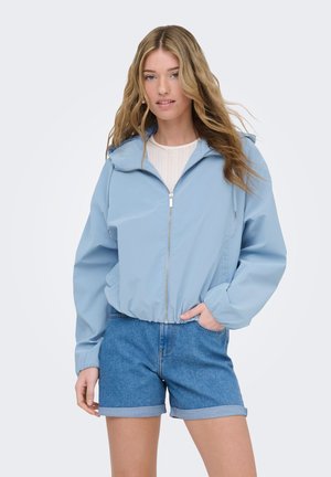 Młoda kobieta o długich blond włosach, ubrana w jasnoniebieską kurtkę z zamkiem i podwinięte szorty dżinsowe, stojąca z jedną ręką w kieszeni.