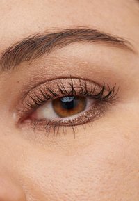 Nærbilde av et øye med mørkebrun iris, lange svarte vipper og metallisk bronse øyeskygge. Huden viser fin tekstur og lette fregner.