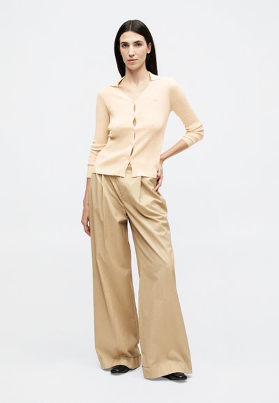 Γυναίκα όρθια με το χέρι στη μέση, φορώντας ένα ροδακινί ριμπ πουκάμισο με κουμπιά και μπεζ παντελόνια φαρδιάς γραμμής με μαύρα παπούτσια.