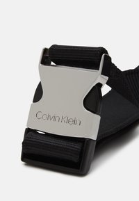 Metallinen Calvin Klein -solki, joka kiinnittää mustan kudotun vyön valkoista taustaa vasten.