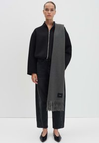 Sciarpa grigia con bordi frangiati, realizzata in materiale morbido, indossata sopra una felpa nera con zip e jeans neri. Scarpe nere flat.