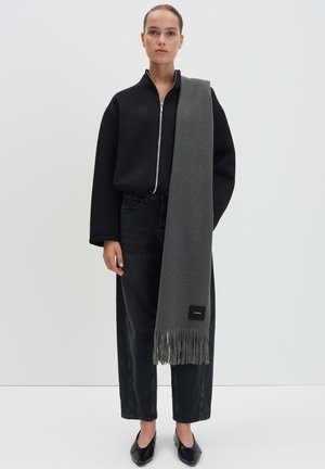 Écharpe grise avec des bords à franges, faite d'un matériau doux, portée sur un sweat à capuche noir zippé et un jean noir. Chaussures plates noires.