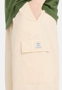 Beiga cargobyxor i en texturerad tyg, med en framficka med en Timberland-logotyp, kombinerat med en grön skjorta.