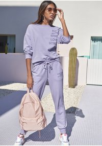 Sudadera y pantalones de chándal a juego en color lila con texto impreso; combinados con una mochila rosa y zapatillas coloridas, en un entorno moderno al aire libre.