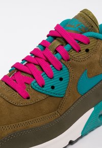 Hnědé semišové tenisky mají jasně růžové tkaničky, tyrkysové akcenty a texturovaný design s výrazným logem Nike swoosh.