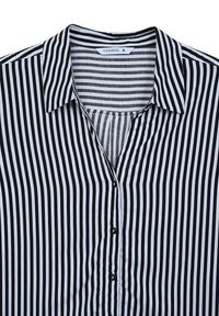 Blusa a righe bianche e nere con collo a camicia, chiusura frontale con bottoni e un tessuto leggero e liscio. Le righe verticali sono uniformi in tutto il capo.