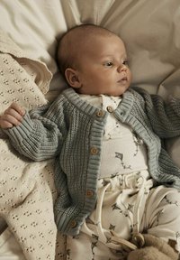 Grauer Strickcardigan mit Holzknöpfen über einem cremefarbenen gerippten Einteiler mit Drachenmuster, auf einem hellen Stoffhintergrund liegend.
