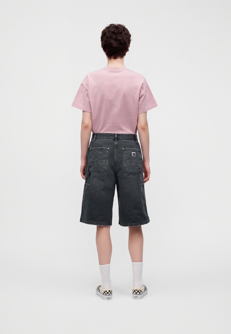 Person steht mit dem Rücken zur Kamera, trägt ein pinkes T-Shirt, dunkelgraue Cargo-Shorts bis zum Knie, weiße Socken und karierte Slip-On-Schuhe.
