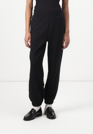 Personne portant un haut sans manches noir côtelé, un pantalon noir avec des poignets élastiques, des chaussettes blanches et des mocassins noirs brillants, debout sur un sol blanc.