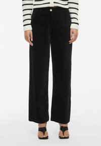 Melnas korduroja plaša kāju bikses ar vertikālu ribu, priekšējo pogu aizdari un sānu kabatām, kopā ar svītrotu džemperi un melnām papēžu kurpēm.