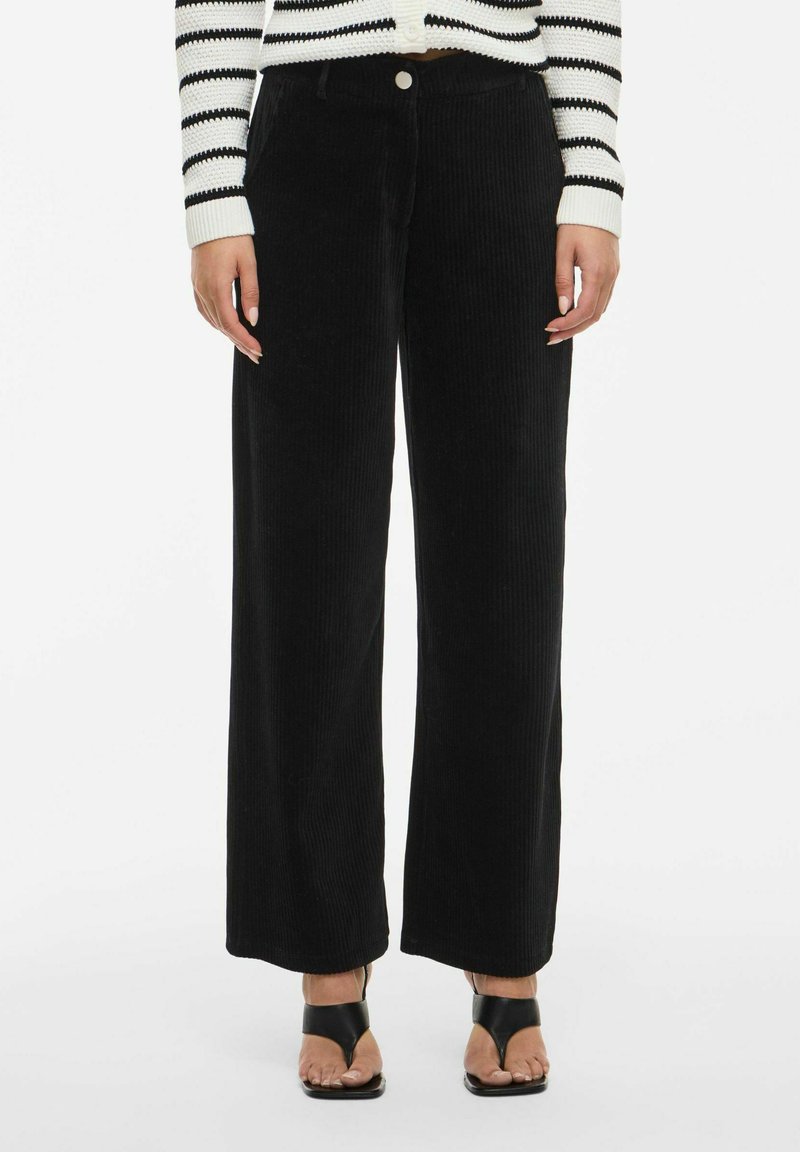 Melnas korduroja plaša kāju bikses ar vertikālu ribu, priekšējo pogu aizdari un sānu kabatām, kopā ar svītrotu džemperi un melnām papēžu kurpēm.