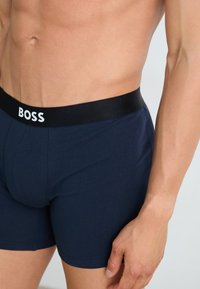 Boxer-briefs bleu marine en tissu doux, avec une taille noire arborant le logo "BOSS" en blanc. Gros plan sur le matériau et la coupe.