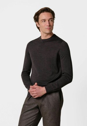 Mann mit dunkelbraunen Haaren, der einen dunklen Langarmpullover und braune Hose trägt, steht mit verschränkten Händen und schaut leicht zur Seite.
