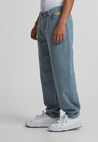 Pantalones de pana de color azul claro con un corte relajado, que cuentan con bolsillos frontales y un logo amarillo en la cinturilla. Combinados con zapatillas blancas.