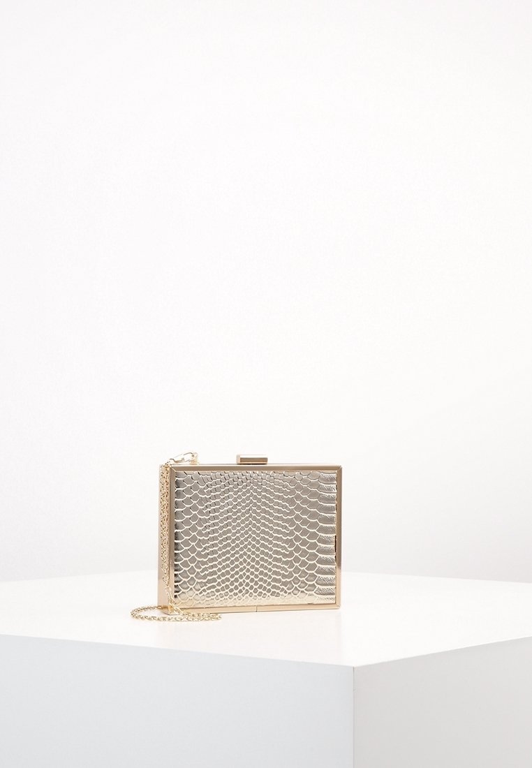 Pochette rectangulaire dorée avec un motif texturé en cuir de serpent et une fine lanière chaîne dorée sur une surface blanche.