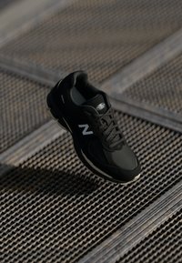 Black athletic shoe with mesh and synthetic materials, featuring a logo on the tongue, laces, and a textured sole, suspended above a grate.