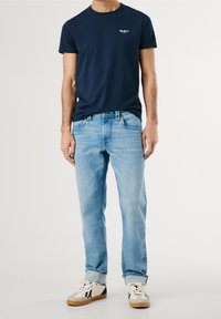 T-shirt in cotone blu navy con logo bianco, abbinata a jeans in denim blu chiaro. Le scarpe hanno una base bianca con accenti neri e suole beige.