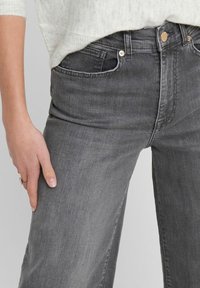 Lähikuva henkilöstä, joka käyttää korkeavyötäröisiä, kuluneita mustia farkkuja ja vaaleaa harmaata neuletta, yksi käsi lepäämällä reidellä.