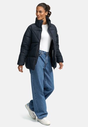 Black puffer jacket with a high collar, quilted design, and contrasting zipper, worn over a white top and paired with loose blue jeans and white sneakers.