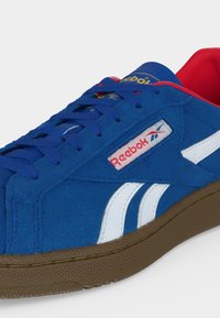 Μπλε αθλητικό παπούτσι Reebok με λευκές πλαϊνές ραβδώσεις, κόκκινη εσωτερική επένδυση, μπλε κορδόνια και καφέ υφασμένη σόλα, εμφανίζεται από εμπρόσθια πλαϊνή γωνία.
