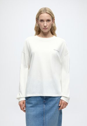 Γυναίκα με μακριά ξανθά μαλλιά φοράει λευκό πουκάμισο με μακριά μανίκια και μικρό κείμενο στο στήθος και μια μπλε τζιν φούστα, στέκεται μπροστά από απλό φόντο.