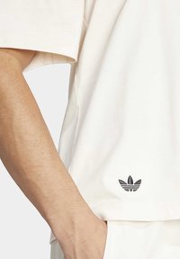 Cream short-sleeve t-shirt made of cotton with a relaxed fit, featuring a small embroidered black logo near the hem. Smooth texture.