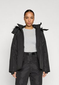 Μαύρο μπουφάν puffer με αποσπώμενη κουκούλα με γούνινη επένδυση, τσέπες με φερμουάρ και λείο ύφασμα, σε συνδυασμό με γκρι πουκάμισο και μαύρο παντελόνι.