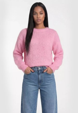 Kobieta z długimi prostymi włosami, ubrana w różowy puszysty sweter i niebieskie dżinsy z wysokim stanem, stojąca z rękami w kieszeniach na białym tle.