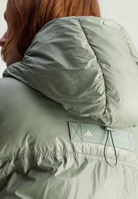Lysegrøn quiltet jakke med hætte, elastisk kant og et logo mærke med korssting detaljer på ryggen. Glat, glansfuld tekstur.